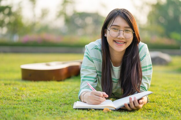 Adolescente con gafas acostado con guitarra y libros en el campo al atardecer.