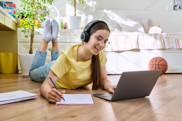 Foto adolescente con gafas estudiando en casa usando una computadora portátil mujer con auriculares y libros de texto tirados en el suelo en la habitación educación adolescencia concepto de escuela secundaria