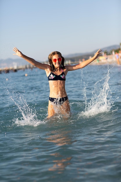 Foto adolescente con gafas de sol rosas divirtiéndose en la playa en el mar jugando con salpicaduras diversión en el concepto de vacaciones de verano