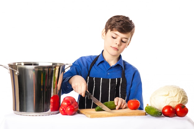 Adolescente con gorro de cocinero cocinero y delantal agrega condimento a la ensaladera en una tabla para cortar, rodeado de verduras y especias aisladas