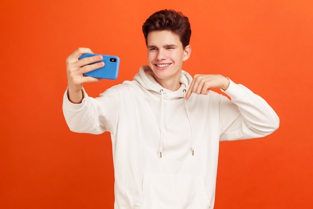 Adolescente guapo sonriente con peinado elegante en capucha casual posando frente al teléfono inteligente, señalando con el dedo haciendo selfie, blogueando. Disparo de estudio interior aislado sobre fondo naranja