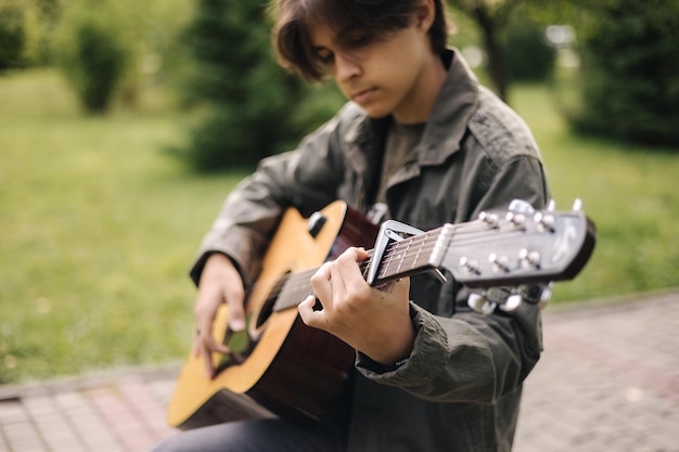 Adolescente guapo tocando la guitarra acústica con capo boy sentado en un banco y tocando música