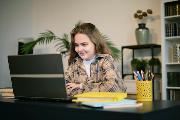 Una adolescente haciendo la tarea en un escritorio en su dormitorio. Una estudiante de la escuela escribe notas. Mira un video. Seminario web en línea. Aprende en una computadora portátil.