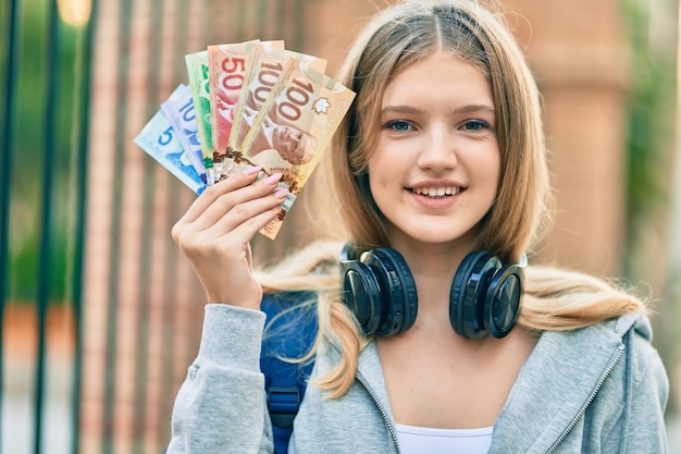 Adolescente hermosa estudiante caucásica usando auriculares con dólares canadienses en la ciudad