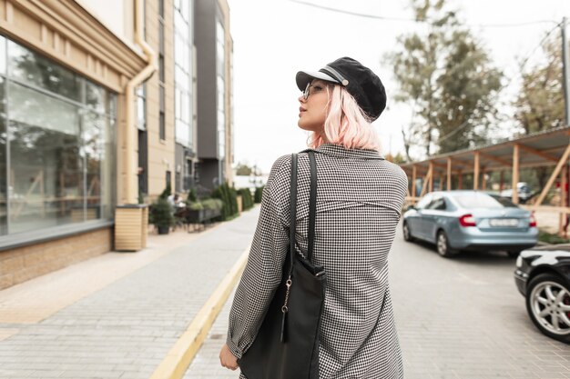 Adolescente hermosa modelo mujer en ropa de moda con gafas vintage y un sombrero con una camisa a cuadros y una bolsa camina por la ciudad