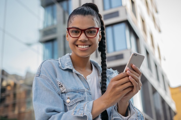 Adolescente hipster sonriente con anteojos elegantes usando una aplicación móvil para comprar en línea al aire libre