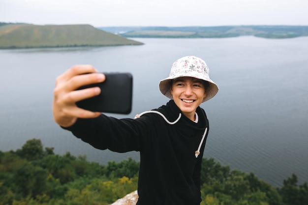 Adolescente hipster tirando selfie no telefone e sorrindo em pé no topo da montanha rochosa com uma vista incrível do rio Jovem estiloso explorando e viajando Momento atmosférico Copie o espaço