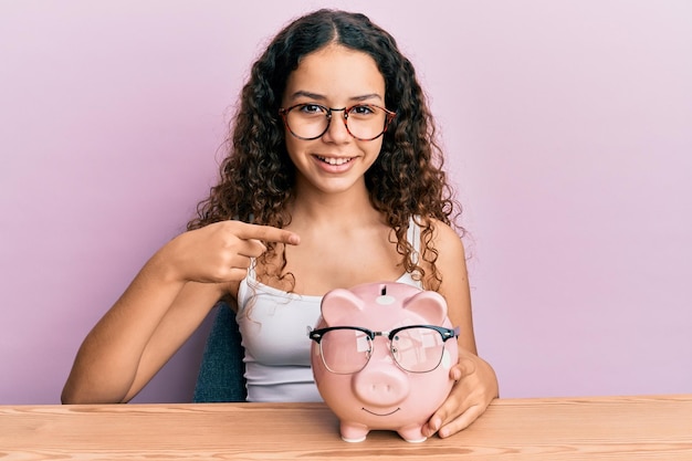 Adolescente hispana sosteniendo alcancía con gafas sonriendo feliz señalando con la mano y el dedo