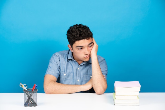 Adolescente hispano aburrido sentado detrás del escritorio aislado sobre fondo azul.