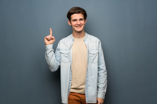 Adolescente, homem, com, jean, jaqueta, sobre, parede cinza, mostrando, e, levantamento, um, dedo, em, sinal, de, a, melhor