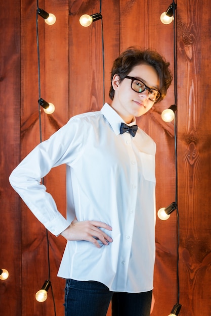 Adolescente inteligente con gafas posando en apartamento tipo loft Adolescente en camisa blanca en bombillas b ...