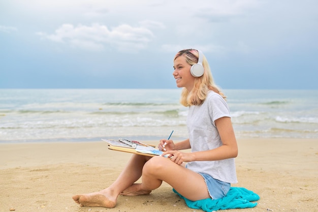 Adolescente jovem em fones de ouvido desenho esboço com aquarelas sentado na praia do mar. Juventude criativa, hobbies de desenho e lazer, talento artístico