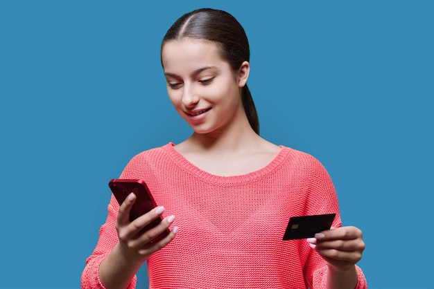 Foto adolescente joven con teléfono inteligente y tarjeta de crédito sobre fondo azul.