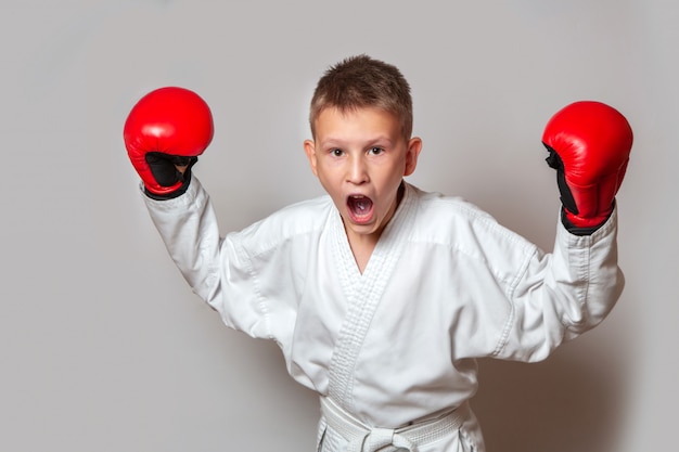 Un adolescente con un kimono blanco y guantes para el combate cuerpo a cuerpo en un gris. Haciendo deportes.