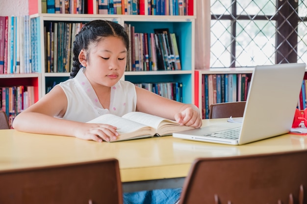 Adolescente lendo o livro com o computador portátil na biblioteca. Conceito de educação e alfabetização