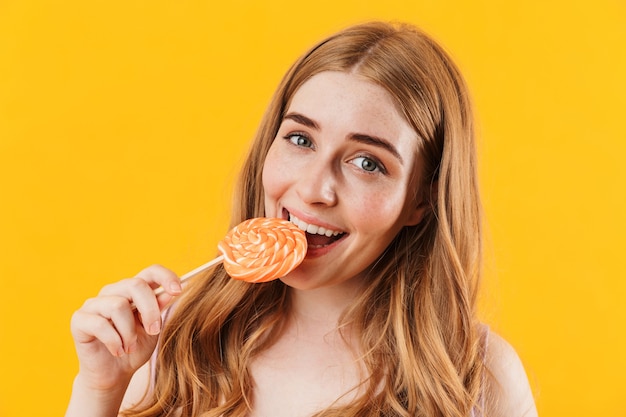 una adolescente linda joven complacida posando aislada sobre pared amarilla con piruleta de caramelo dulce.