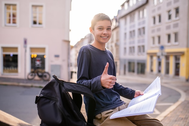 Adolescente lindo alegre sentado al aire libre con un libro de texto abierto en la mano haciendo un signo de aprobación