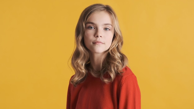 Adolescente loira atraente de cabelos ondulados em suéter vermelho parecendo confiante posando na câmera isolada em fundo amarelo