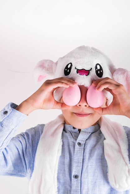 Foto adolescente en una máscara de conejito de pascua cierra los ojos con huevos pintados a mano de color rosa sobre fondo gris claro.