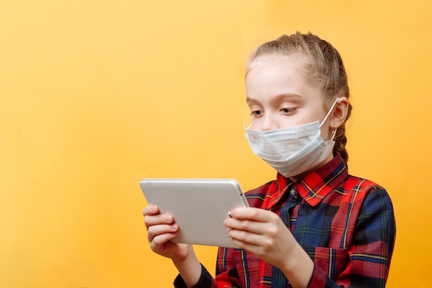 Una adolescente con una máscara médica en una pared amarilla sostiene una Tablet PC