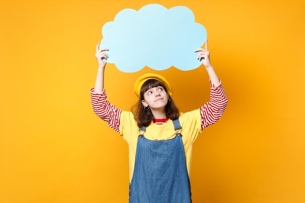 Adolescente menina bonita pensativa na boina francesa, vestido jeans olhando para cima em branco vazio azul Diga nuvem, bolha do discurso isolada em fundo amarelo. Conceito de estilo de vida de emoções de pessoas. Mock up espaço de cópia.