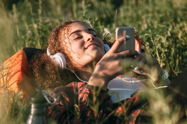 Foto adolescente con mochila acostada en un prado escuchando música con auriculares y teléfono celular