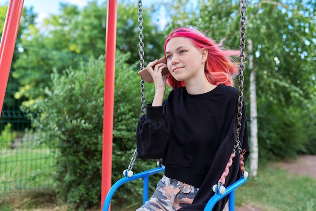 Adolescente de moda con el pelo teñido con un smartphone montado en un columpio, escuchando un mensaje de audio. Estilo de vida de los adolescentes, comunicación y tecnología, concepto de juventud.