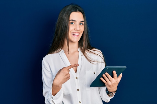 Foto adolescente morena joven que usa el dispositivo del panel táctil que sonríe feliz que señala con la mano y el dedo