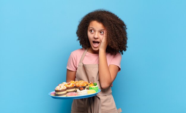 Adolescente muito afro se sentindo feliz, animado e surpreso, olhando para o lado com as duas mãos no rosto. conceito de padeiro humorístico