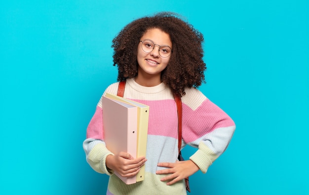 Adolescente muito afro, sorrindo alegremente com uma mão no quadril e uma atitude confiante, positiva, orgulhosa e amigável. conceito de estudante