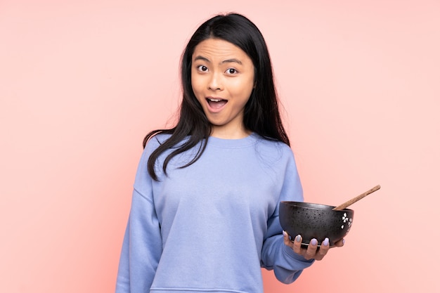 Adolescente mujer asiática en la pared de color beige con sorpresa y expresión facial sorprendida mientras sostiene un tazón de fideos con palillos