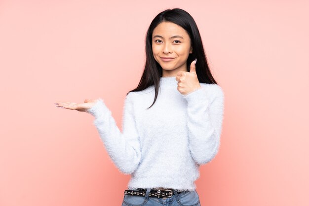 Adolescente mujer china aislada en rosa sosteniendo copia espacio imaginario en la palma para insertar un anuncio y con los pulgares hacia arriba