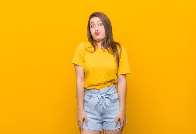 Adolescente mujer joven con una camisa amarilla sopla las mejillas, tiene expresión cansada. Concepto de expresión facial.