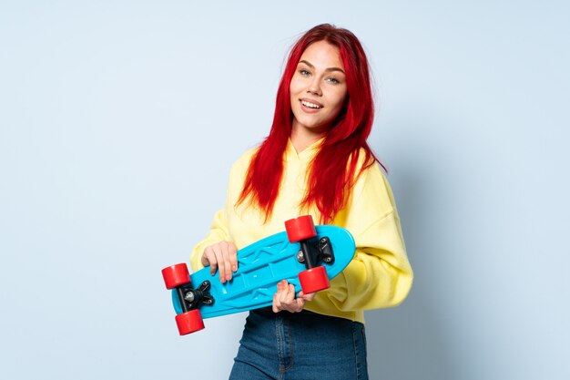 Adolescente mujer de pelo rojo aislada en la pared azul con un patín con expresión feliz