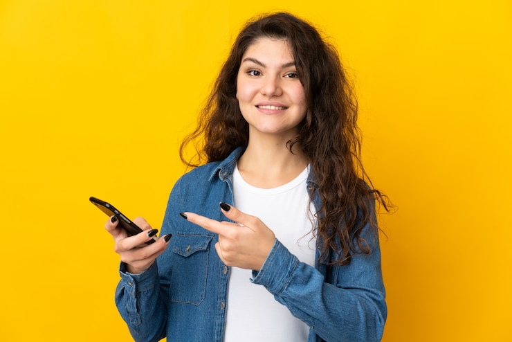 Foto adolescente mujer rusa aislada en la pared amarilla mediante teléfono móvil y apuntando