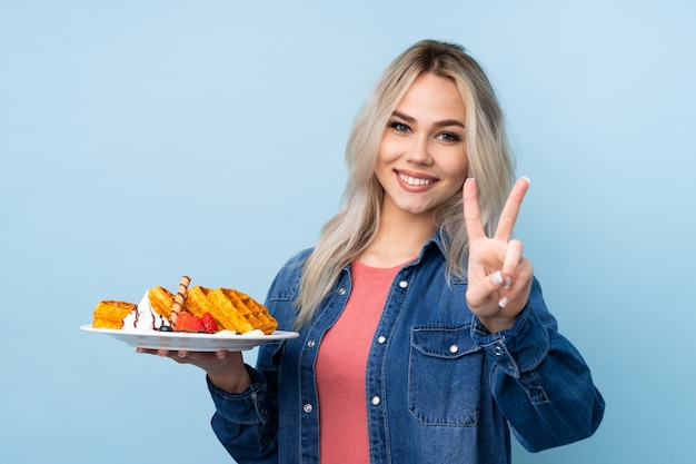 Adolescente mujer sosteniendo waffles sobre pared azul aislado sonriendo y mostrando el signo de la victoria