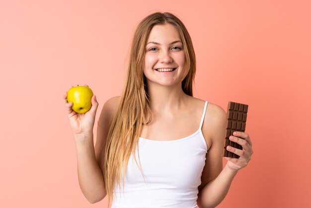 Adolescente mujer ucraniana aislada en el espacio rosa con una tableta de chocolate en una mano y una manzana en la otra