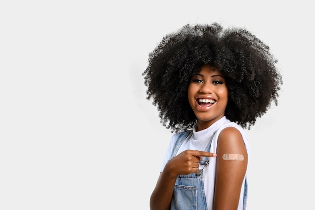 Foto adolescente negra aponta para adesivo em seu braço mostrando que ela foi vacinada, isolada em fundo cinza.