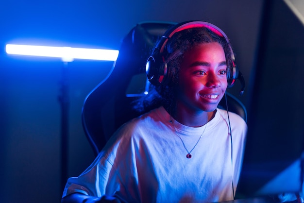 Adolescente negra sonriente con auriculares jugando videojuegos en un club de videojuegos con iluminación azul y roja