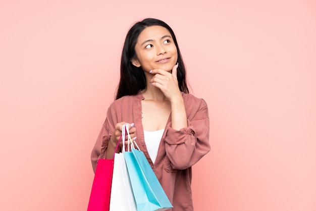 Adolescente niña china aislada en la pared rosa sosteniendo bolsas de compras y pensando