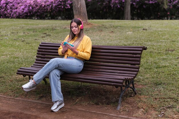 Foto adolescente ouvindo música com seus fones de ouvido conectados a um telefone celular em um banco do parque