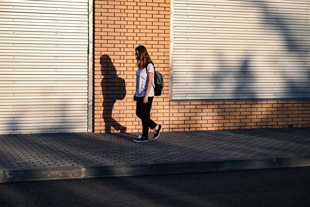 Una adolescente con pantalones negros y una camiseta blanca camina por la calle con una mochila negra en la espalda de vuelta a la escuela