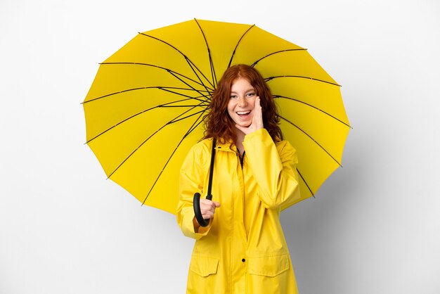 Adolescente pelirroja abrigo impermeable y paraguas aislado sobre fondo blanco con sorpresa y expresión facial conmocionada