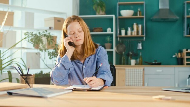 Foto adolescente pelirroja estudiando en casa en línea hablando por teléfono