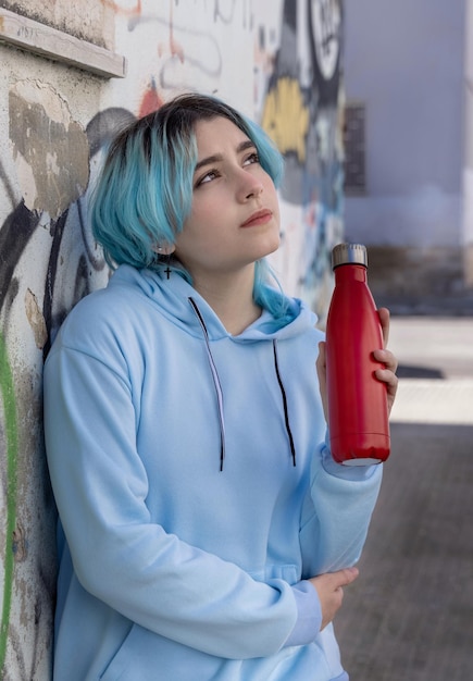 Adolescente de pelo azul con capucha azul que se queda cerca de la pared de graffiti con una botella de agua roja