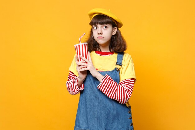 Adolescente pensativa con boina francesa, vestido de mezclilla sosteniendo una taza de plástico de cola o soda aislada en el fondo de la pared amarilla en el estudio. Emociones sinceras de la gente, concepto de estilo de vida. Simulacros de espacio de copia.