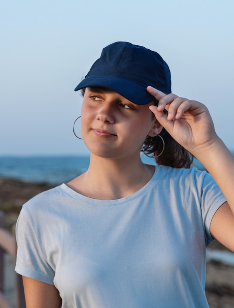 Adolescente de pie junto al mar al atardecer. Jovencita vestida con camiseta y gorra de béisbol azul oscuro y tocando la visera. Maqueta de gorra y camiseta