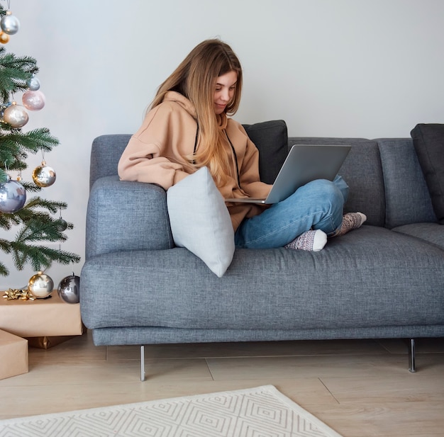 Adolescente con portátil sentado en un sofá en la sala de estar cerca del árbol de Navidad