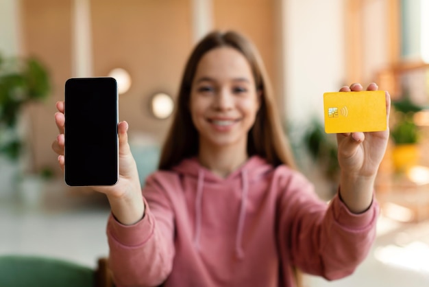 Foto adolescente positiva mostrando teléfono inteligente con pantalla en blanco y tarjeta de crédito sentada en casa maqueta de enfoque selectivo
