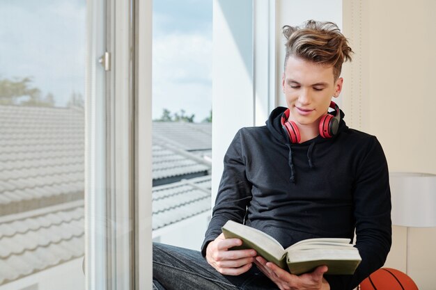 Adolescente positivo sentado en la ventana de su habitación y leyendo un nuevo libro cautivador